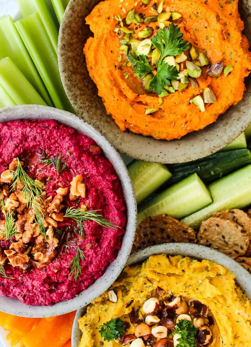 Three dips photographed from above — roasted red pepper with pistachios, beetroot with walnuts and dill, and a hummus with hazelnuts — with celery, cucumber and seeded crackers around them