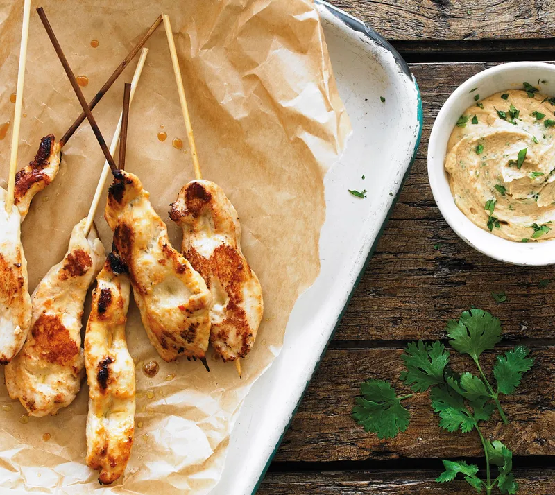 Grilled chicken skewers on a lined tray beside a bowl of hummus, with fresh cilantro on the wood beside it