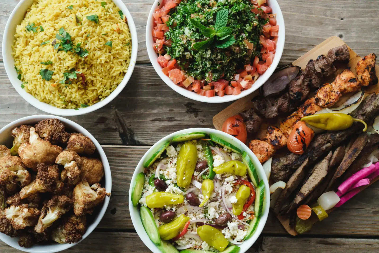 An overhead spread on a wood table — saffron rice, tabbouleh, fried cauliflower, a greek salad with olives and pepperoncini, and chicken and beef skewers