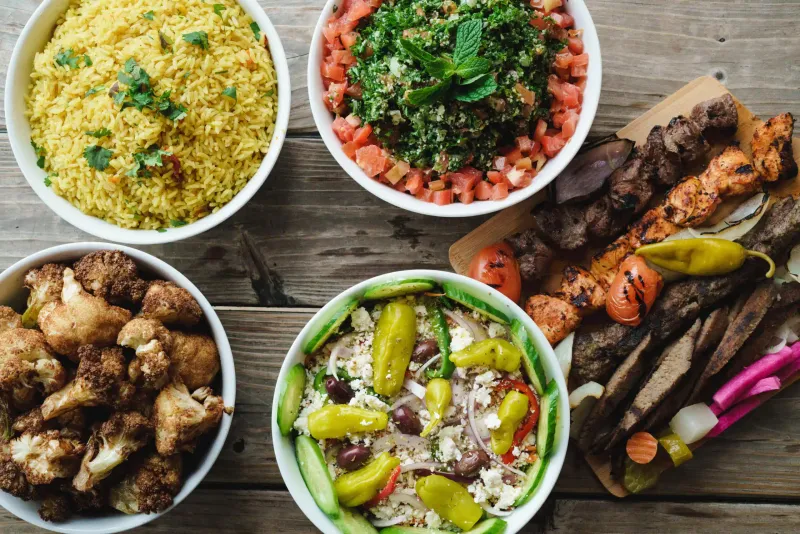 An overhead spread on a wood table — saffron rice, tabbouleh, fried cauliflower, a greek salad with olives and pepperoncini, and chicken and beef skewers