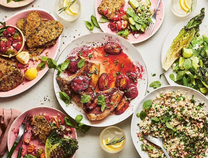 A laid table of plated mains — herb-crusted fish, roasted plums, charred greens and a herbed grain salad — on pink and cream plates
